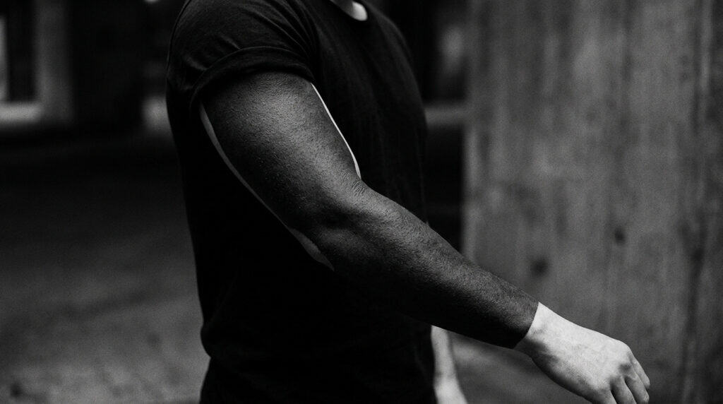 Close-up of a man's muscular arm with a sleek, athletic appearance, wearing a black shirt, in a black-and-white photo emphasizing strength and fitness.