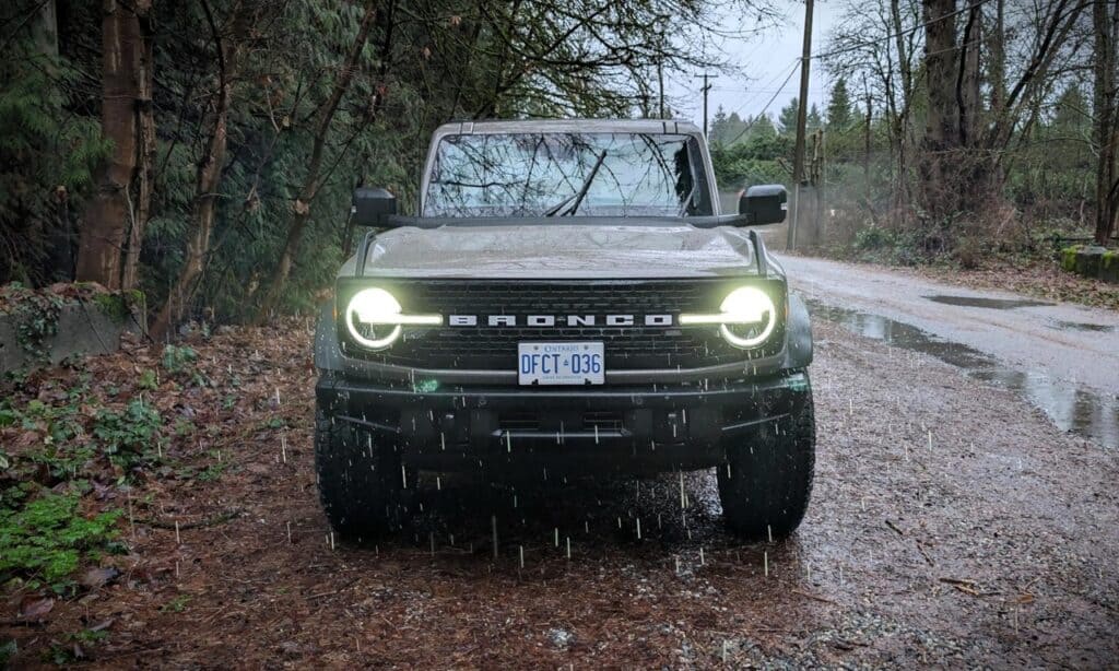 Front view of a 2026 Ford Bronco Badlands 4-Door SUV driving through a muddy forest trail, showcasing rugged off-road capabilities and modern design features.