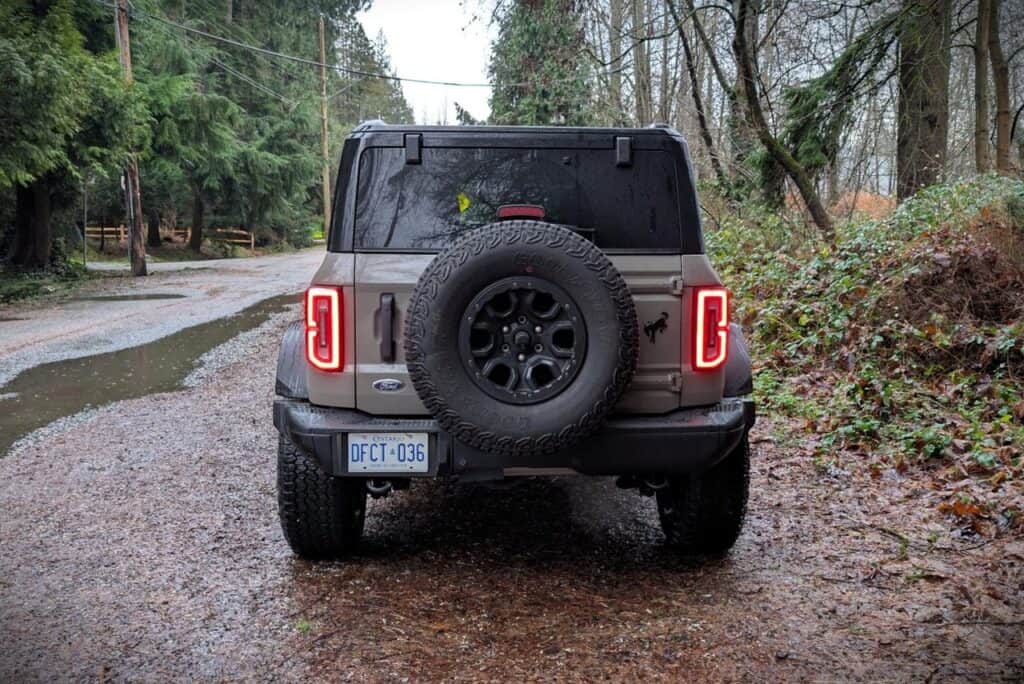 A detailed view of the rear of a 2026 Ford Bronco Badlands 4-Door SUV parked on a muddy forest trail, showcasing its rugged design, spare tire, and off-road capabilities.