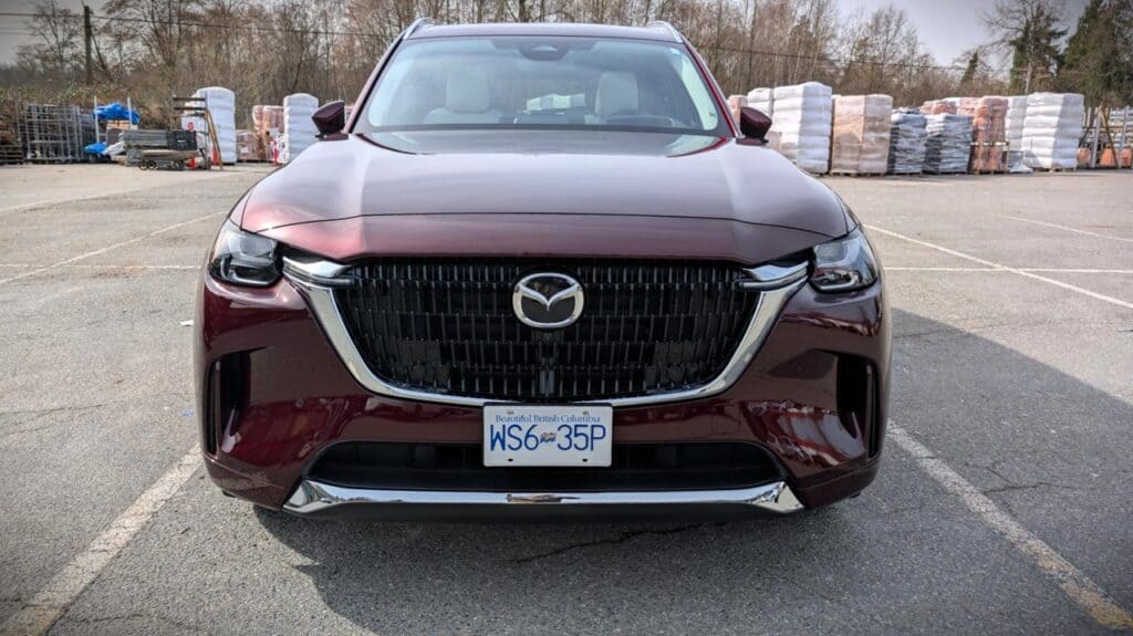 2026 Mazda CX-90 PHEV Signature Review 2 Modern maroon Mazda CX-90 PHEV SUV parked outdoors in a lot with trees and stacked materials in the background, showcasing sleek design and front grille details.