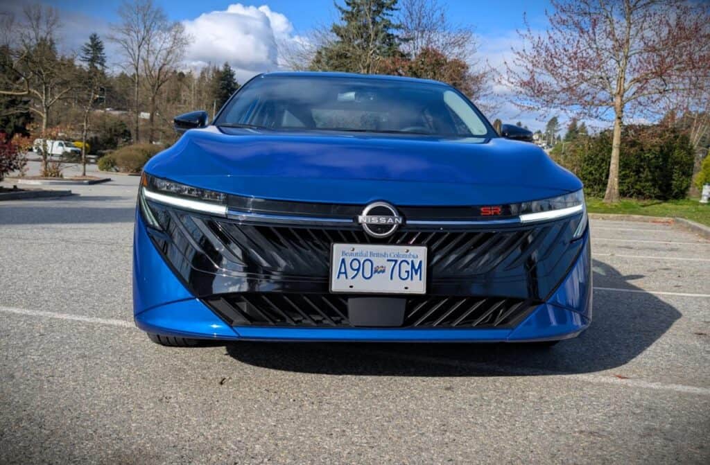 A sleek blue Nissan electric vehicle parked in an outdoor lot with trees and a partly cloudy sky in the background.