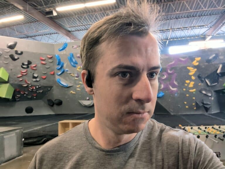 Wireless earbuds worn by a man at an indoor bouldering gym with colorful climbing holds in the background.