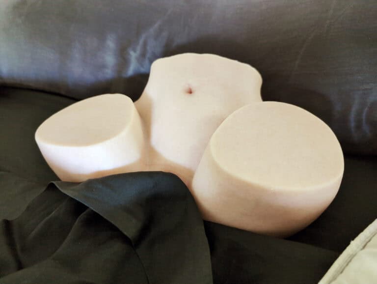 A woman’s torso sculpture made of pinkish stone, featuring a small belly button, with two large rounded stone pieces on either side, placed on a dark fabric surface, creating an artistic and modern ae.