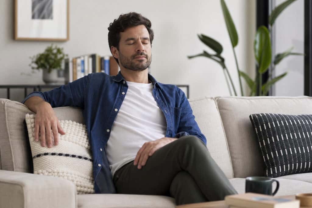 Stressed Out Man: Why Trying to 'Fix' Him Always Backfires 5 Man sitting on sofa with eyes closed, practicing meditation or mindfulness in a modern living room setting.