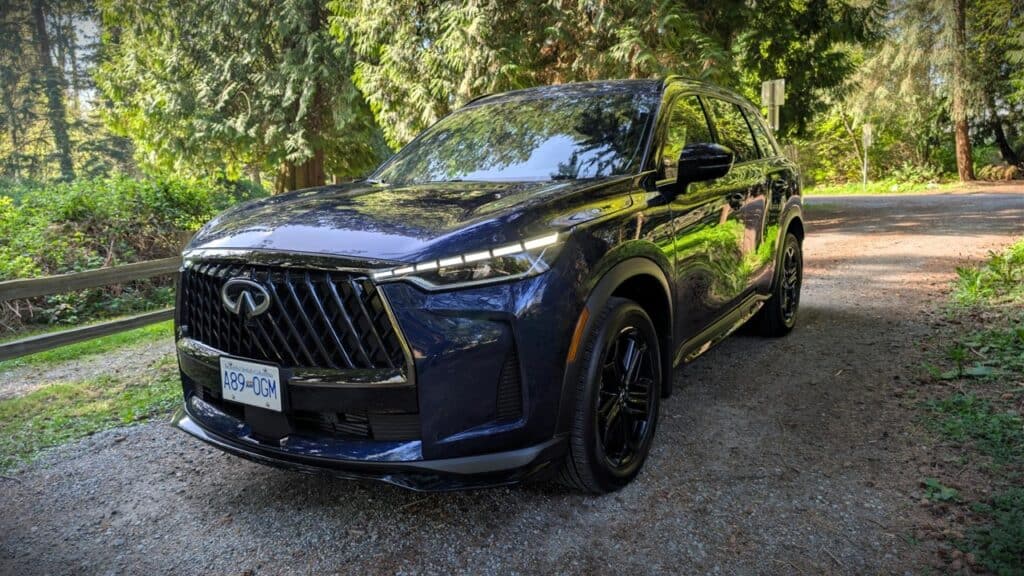 2026 Infiniti QX60 Sport Review 6 2026 Infiniti QX60 Sport in deep blue parked on a forested road with lush greenery and tall trees in the background.