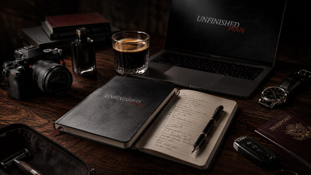 Desk setup with Unfinished Man branding, including a laptop, camera, notebook, pen, glass of whiskey, and travel accessories.