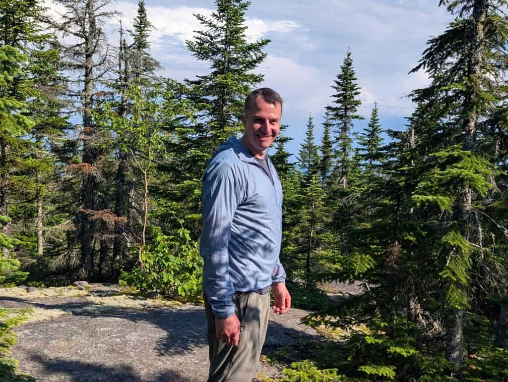 Man hiking in a forested area with tall pine trees and a mountain view in the background.