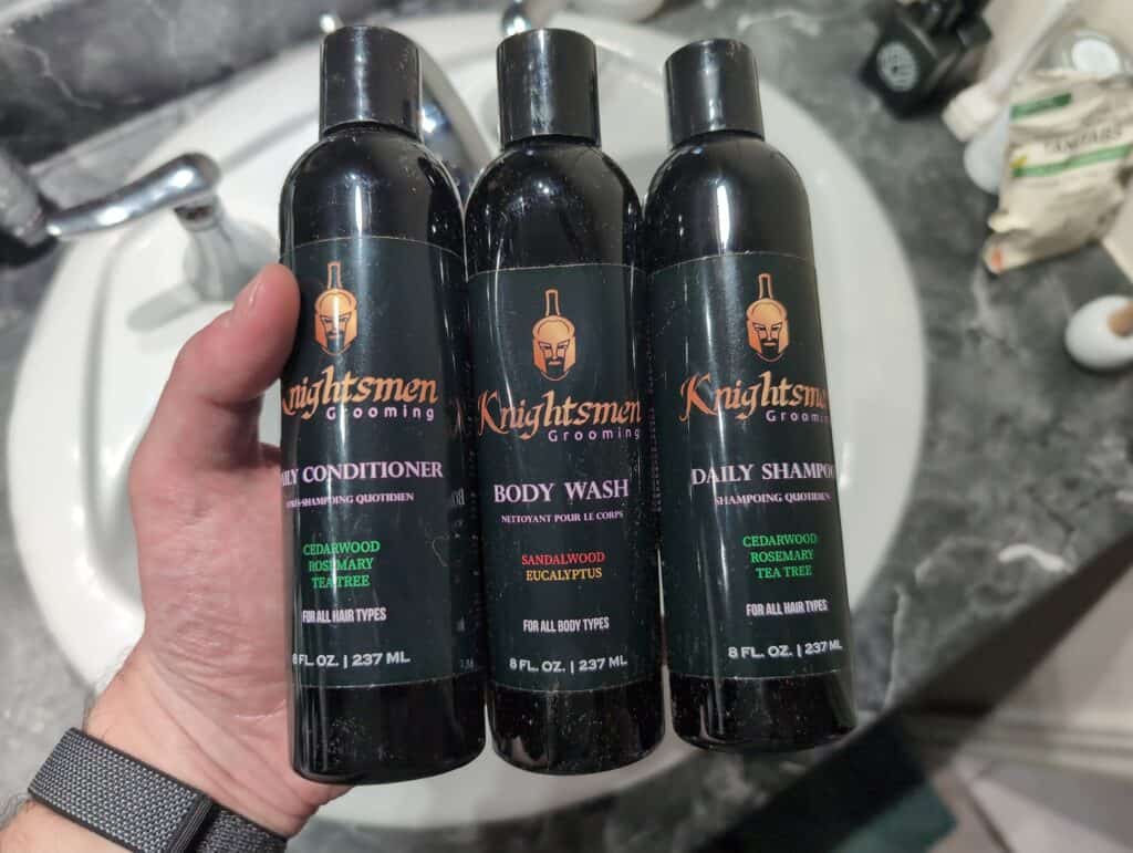 Knightsmen Review: Is It the Best Rosemary Shampoo and Conditioner? 2 A woman’s hand holding three black bottles of Knightsmen Grooming products, including conditioner, body wash, and shampoo, on a bathroom countertop with various toiletries in the background.