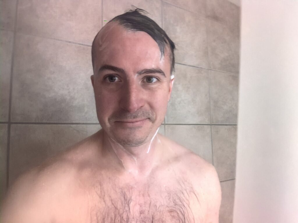 Knightsmen Review: Is It the Best Rosemary Shampoo and Conditioner? 3 Relaxed man taking a shower, smiling, with wet hair and bare shoulders, in a modern tiled bathroom.