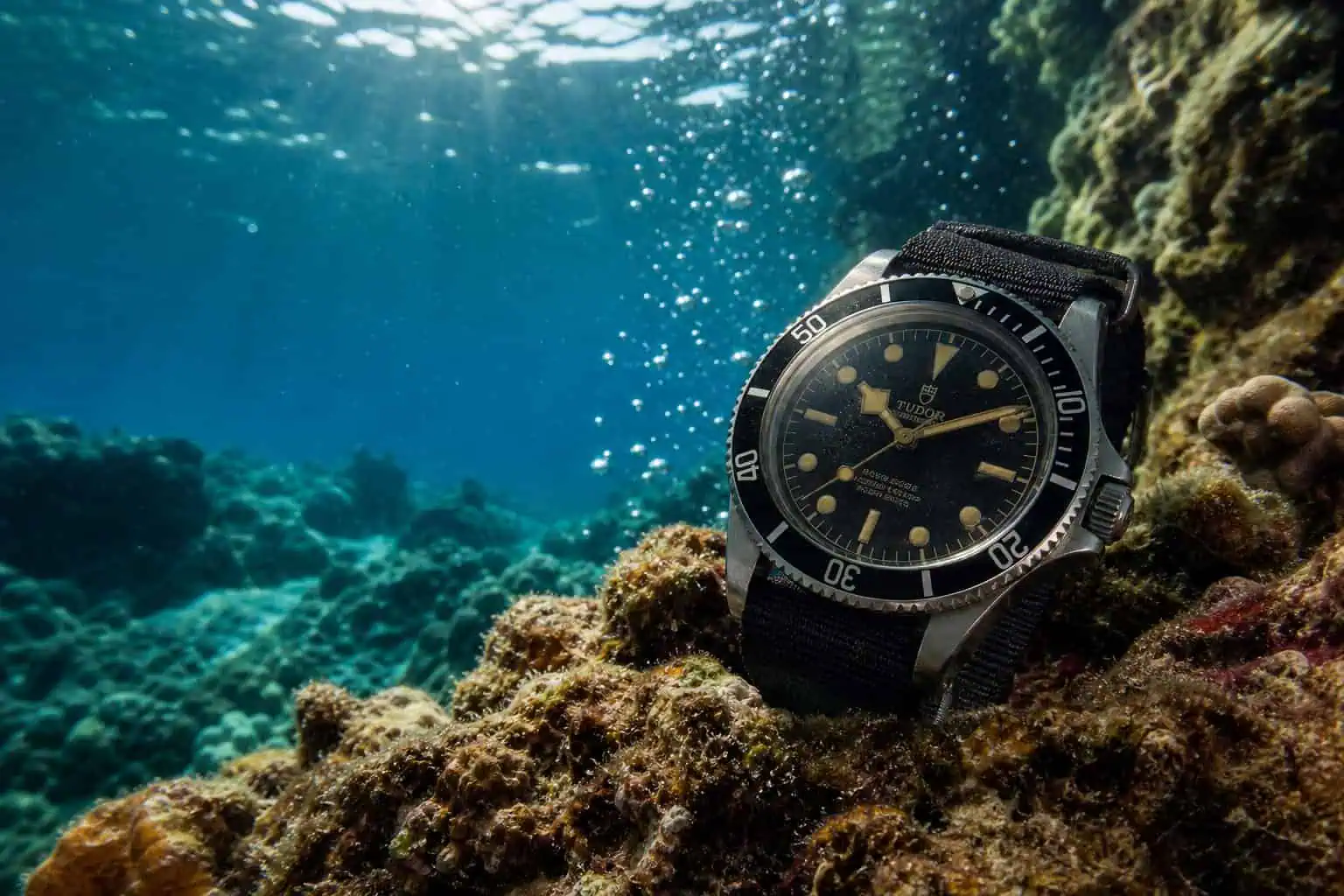 Vintage dive watch on a coral reef underwater with sunlight rays and bubbles.