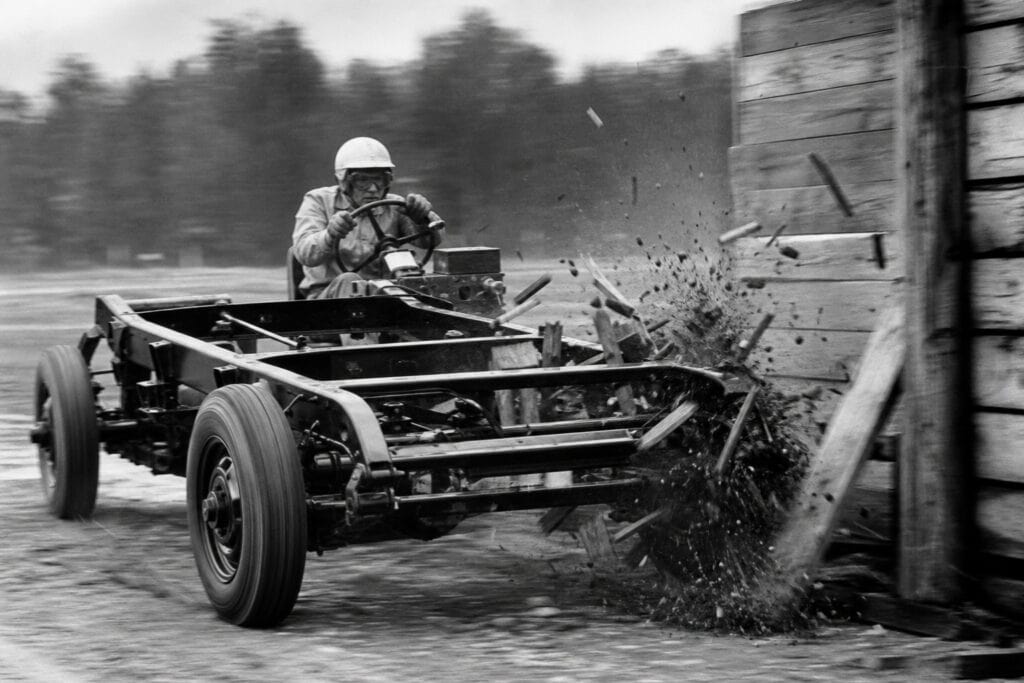 Black and white image of a military vehicle crashing into a wooden barrier during a training exercise, with a soldier operating the vehicle and debris flying.