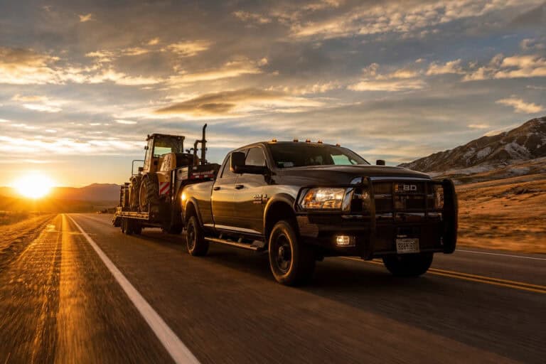 A black heavy-duty pickup truck towing a tractor trailer on an open road during sunset, with mountains in the background.