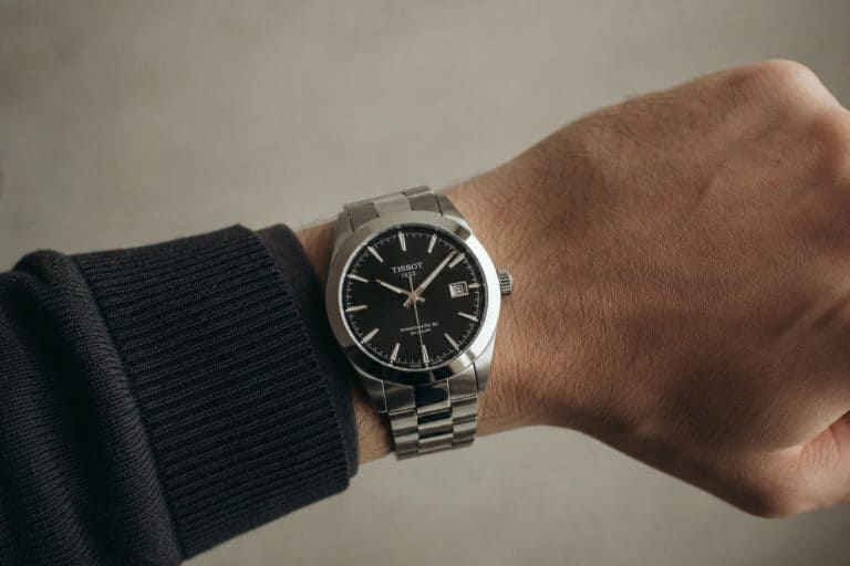Close-up of a men's silver wristwatch with black dial and date display, worn on a person's wrist with a black sleeve, against a neutral background.