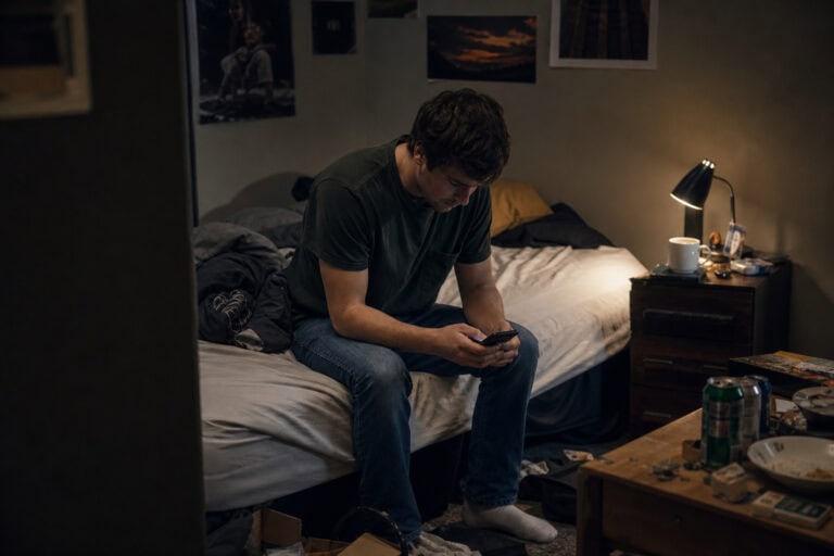 Young man sitting on bed in a dimly lit bedroom, focused on his smartphone, with a bedside lamp and cluttered table nearby.