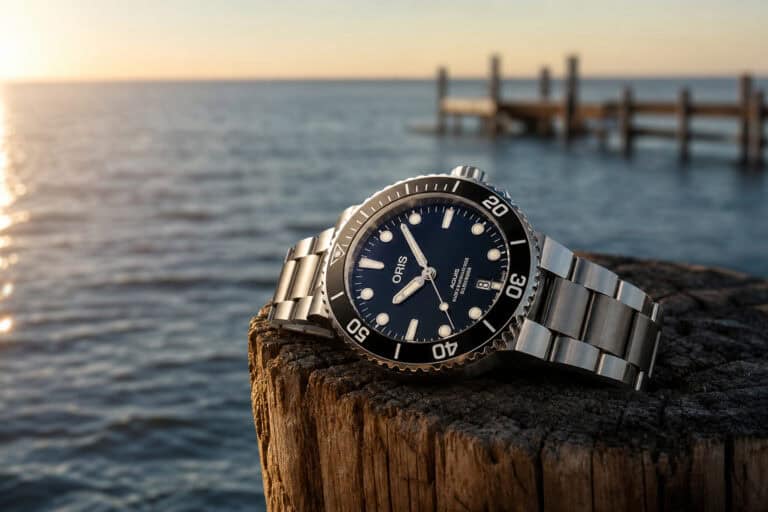 Luxury Oris dive watch with black dial and stainless steel bracelet resting on a wooden post by the water at sunset.