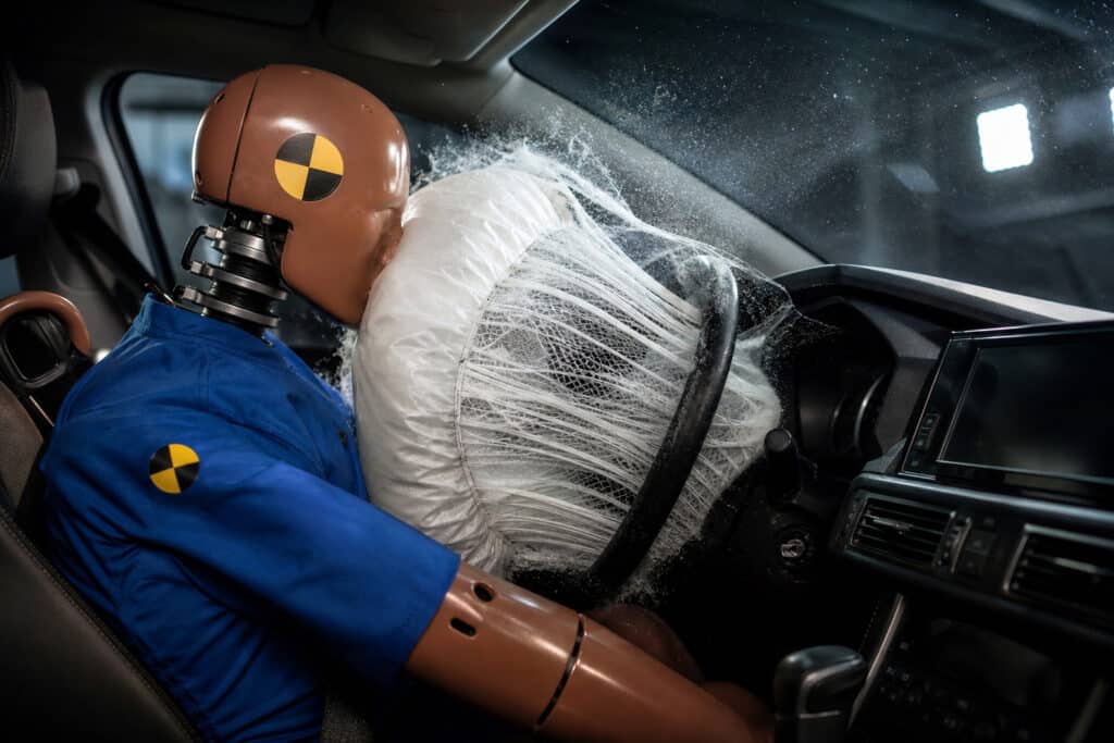 Crash test dummy in blue suit hitting the steering wheel during a simulated car crash impact test inside a vehicle.