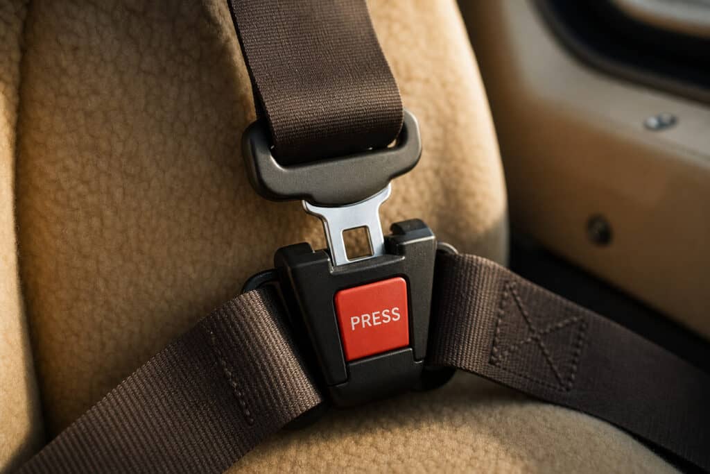 Close-up of a car seat belt buckle with a red "PRESS" button, secured to a beige car seat, emphasizing vehicle safety and seat belt importance.