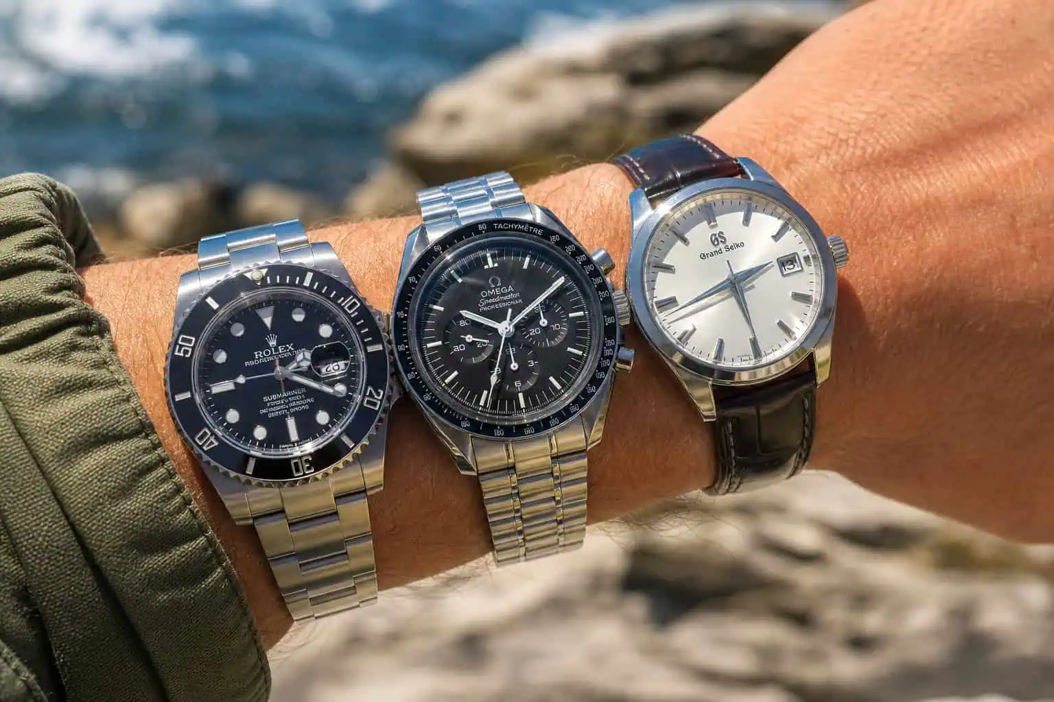 Three luxury wristwatches displayed on a person's arm outdoors near water, showcasing high-end design and craftsmanship.