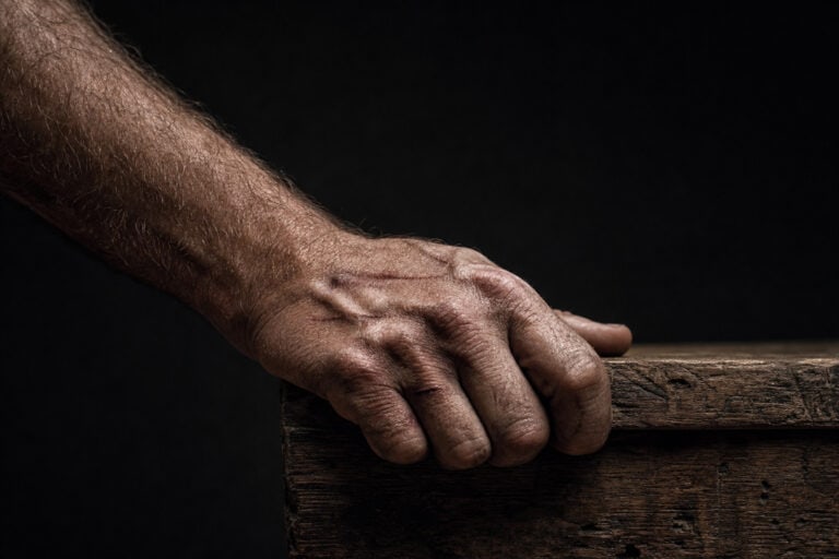 A rugged, weathered hand firmly grasps the edge of a wooden surface, highlighting texture and strength against a dark background.