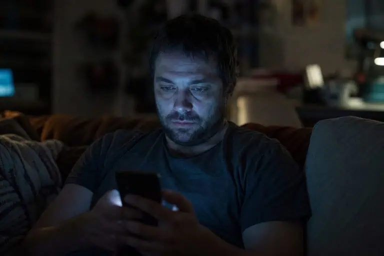 A man with dark hair and beard is focused on his smartphone in a dimly lit room, illuminated by the screen's glow, creating a moody and contemplative atmosphere.