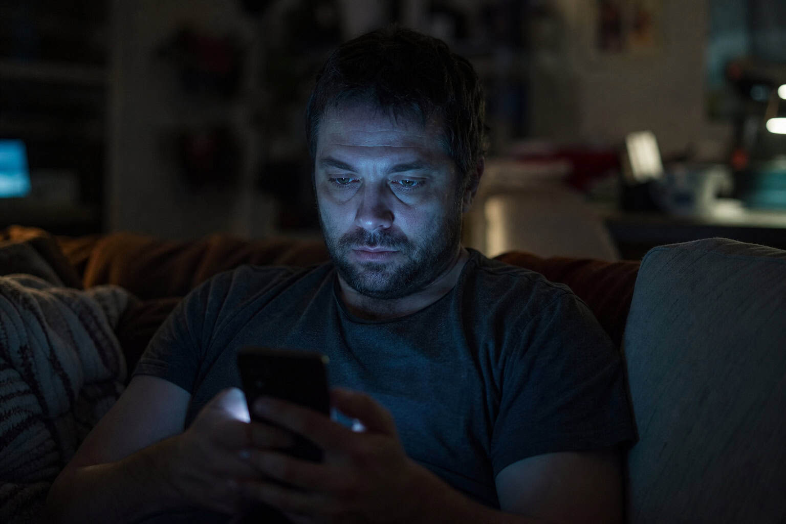 A man with dark hair and beard is focused on his smartphone in a dimly lit room, illuminated by the screen's glow, creating a moody and contemplative atmosphere.