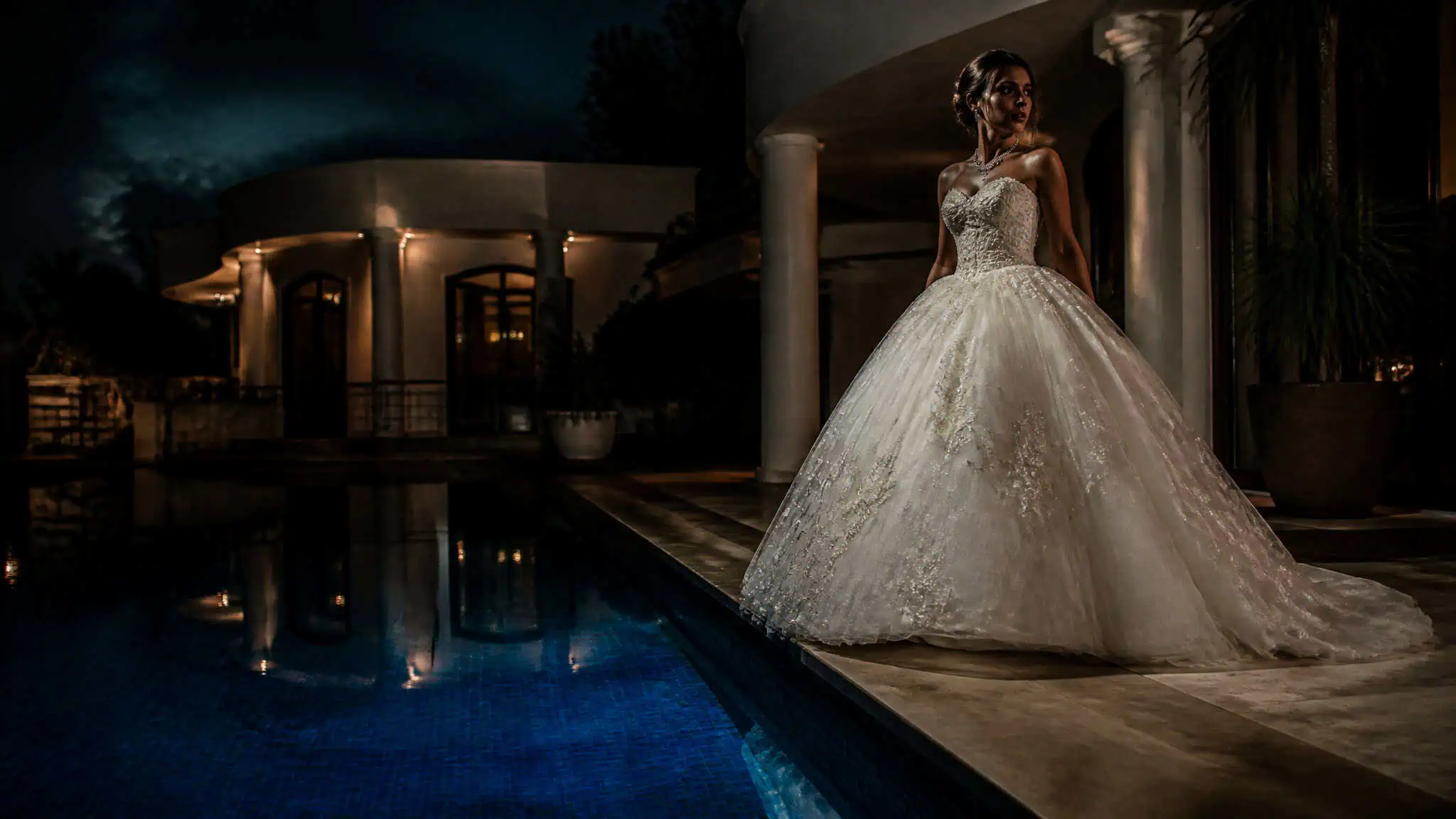 Bride in heavy ballgown standing near pool edge at night