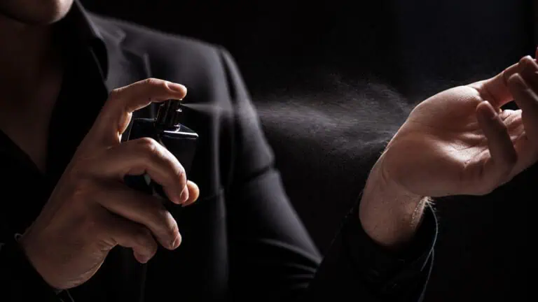 A man in a dark suit sprays perfume from a black bottle, with a fine mist visible in the air, creating a sophisticated and elegant atmosphere.