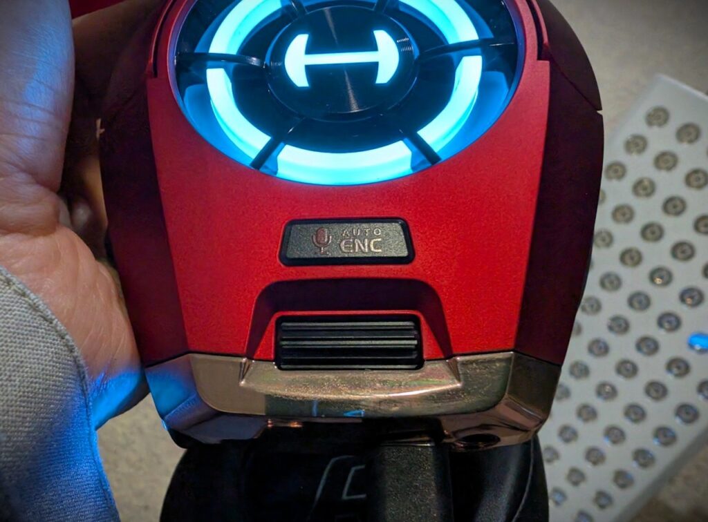 Edifier GX Professional Gaming Headset Review: Fortnite Nights With My Son Settled It 2 Close-up of a red gaming headset with a blue LED ring and a labeled CNC switch, held by a person, against a blurred perforated metal background.