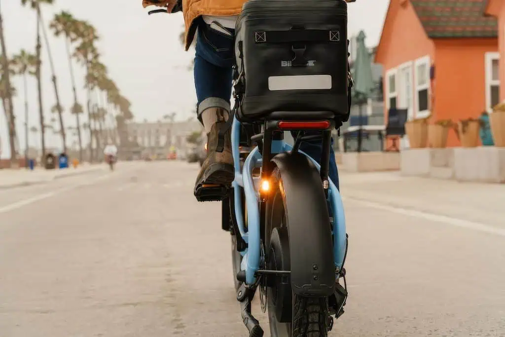 Segway MUXI Pro Cargo e-Bike: Full Specs, $1,999.99 Price, and the Fine Print 4 Close-up of a person riding a blue Segway MUXI Pro Cargo e-Bike on a city street with palm trees and colorful buildings in the background.