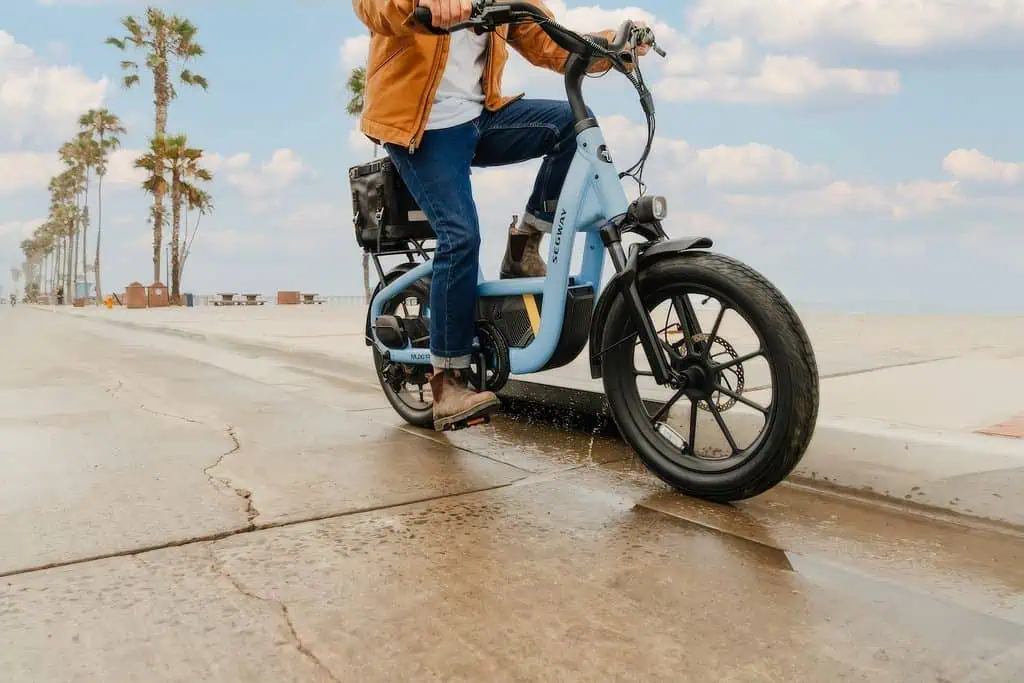 Segway MUXI Pro Cargo e-Bike: Full Specs, $1,999.99 Price, and the Fine Print 2 Person riding a Segway MUXI Pro Cargo e-Bike along a beachside promenade with palm trees and a cloudy sky.