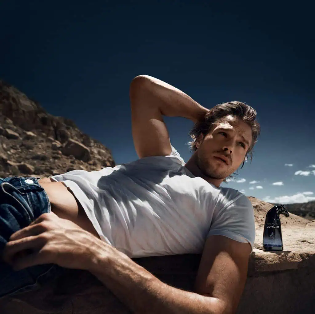 Stetson Untamed Fragrance Collection: Full Notes for Open Sky, Wild Sage, and Free Rein 3 Male model lying on a rock in a desert landscape with a fragrance bottle nearby, under a clear blue sky.