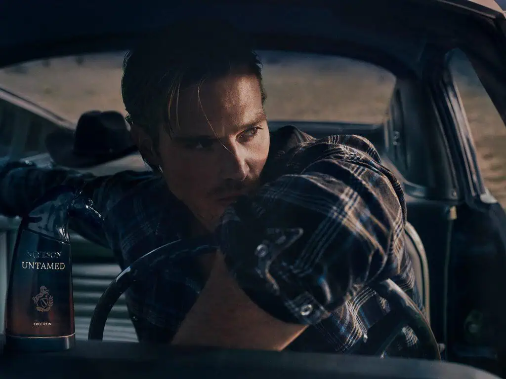 Stetson Untamed Fragrance Collection: Full Notes for Open Sky, Wild Sage, and Free Rein 4 A young man with styled hair and a plaid shirt sitting in a vintage car, looking contemplative with a bottle of Stetson Untamed fragrance nearby.