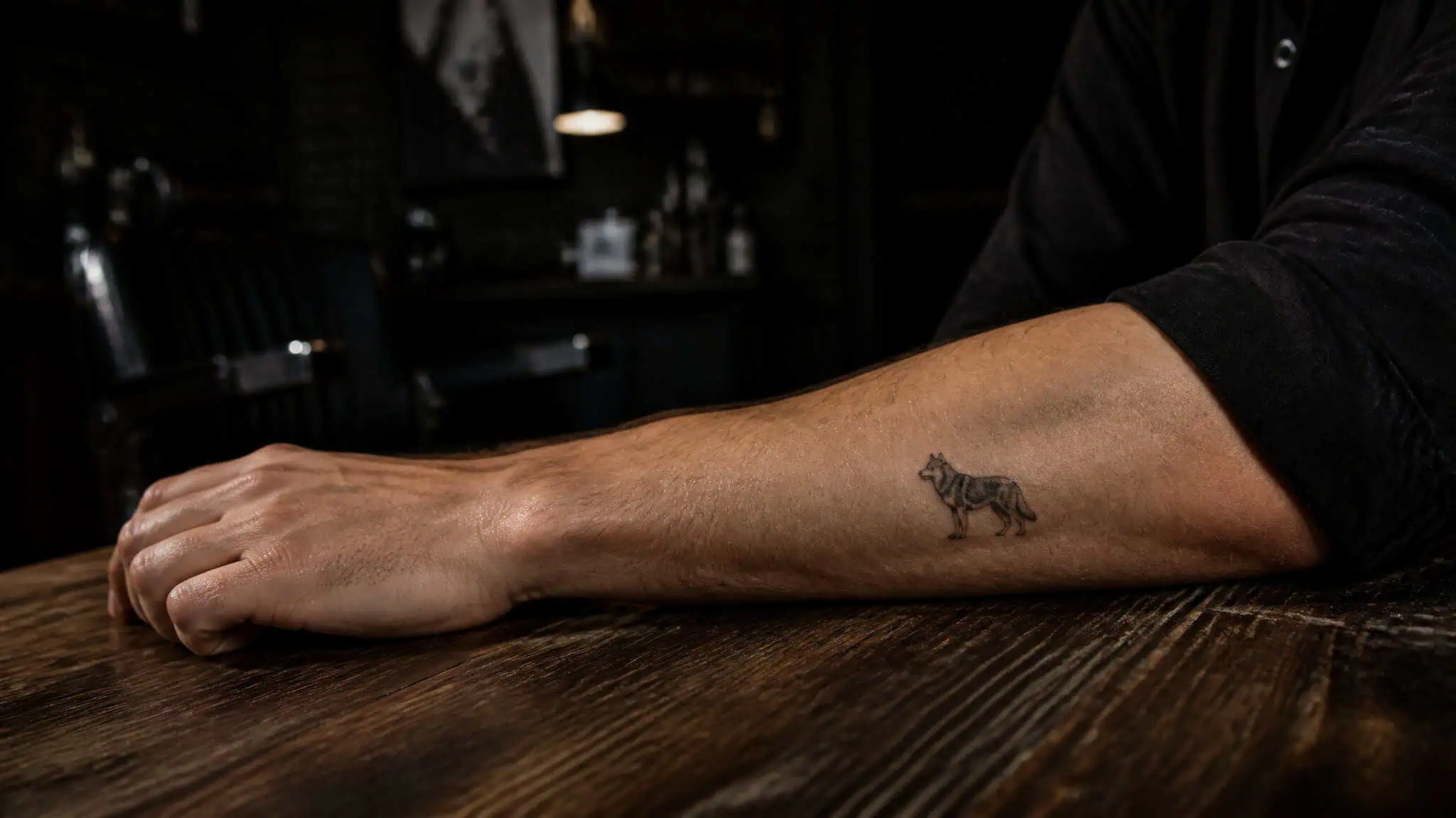 Close-up of a man's arm with a small tiger tattoo resting on a dark wooden bar counter in a dimly lit bar setting.