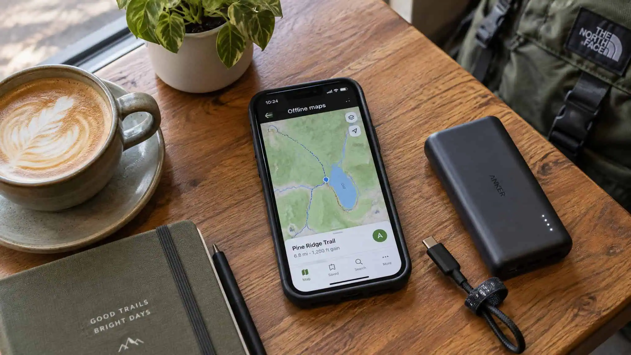 Best Travel Hacks for Women: Pickpocketing, Shipwreck, and the Systems That Actually Work 3 Smartphone displaying a hiking trail map on OpenRidge Trail with nearby lake, placed on a wooden table alongside a coffee cup, portable charger, and a notebook.
