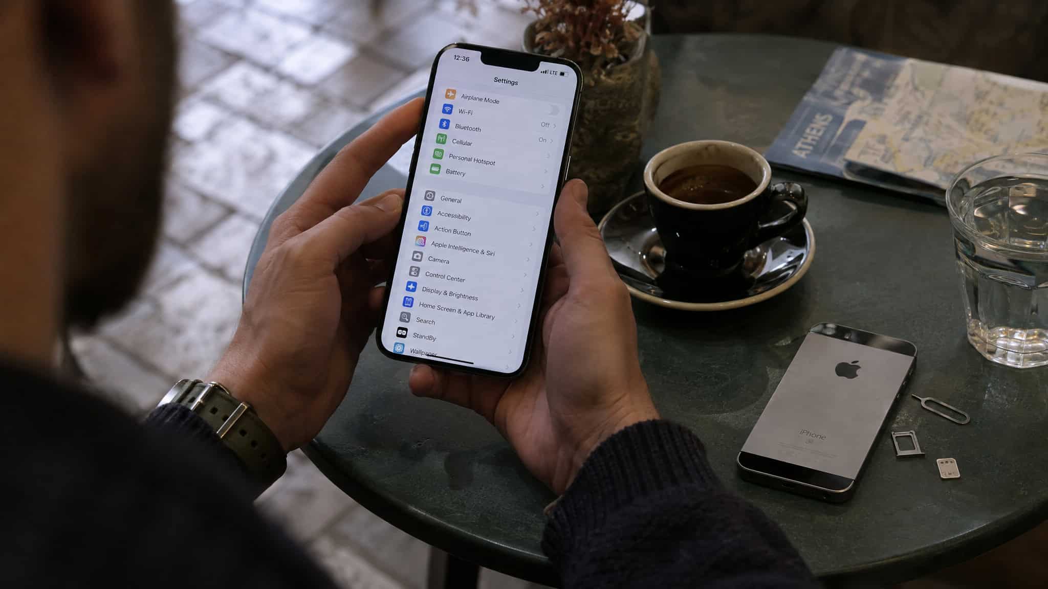 Is eSIM Good for International Travel? Pros and Cons After 3 Years of Testing 4 Close-up of a person holding a smartphone displaying app icons, with a coffee cup, iPhone, and glass of water on a cafe table.