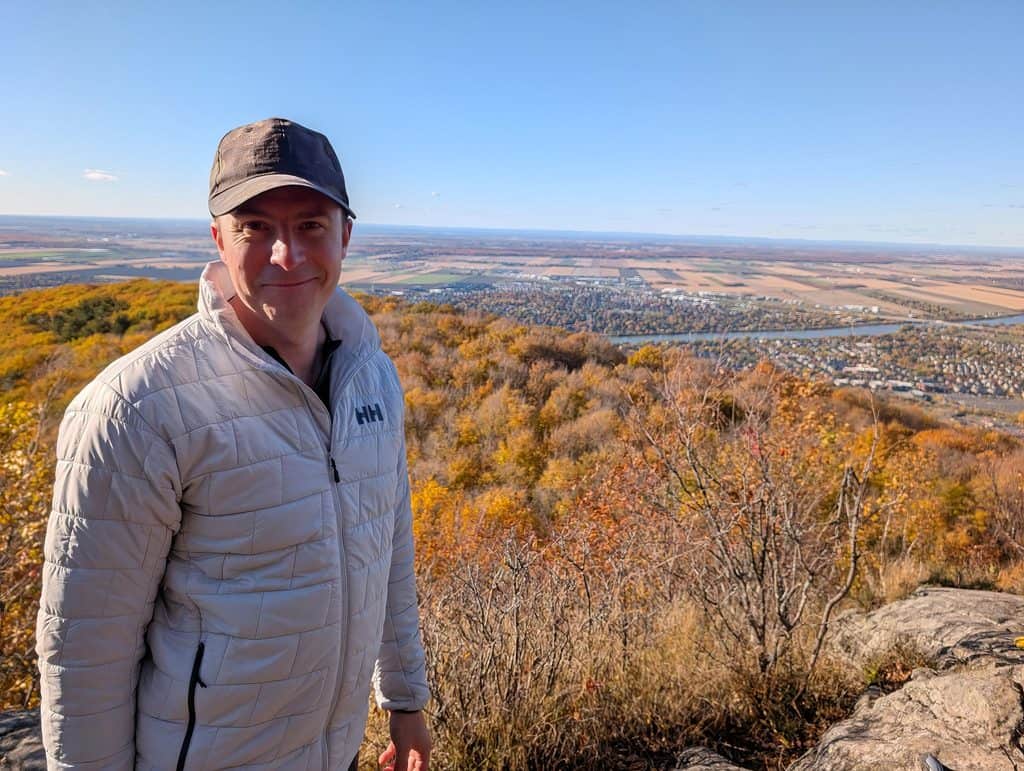 Man in light gray Helly Hansen jacket and black cap standing on a hilltop with autumn trees and a distant landscape.