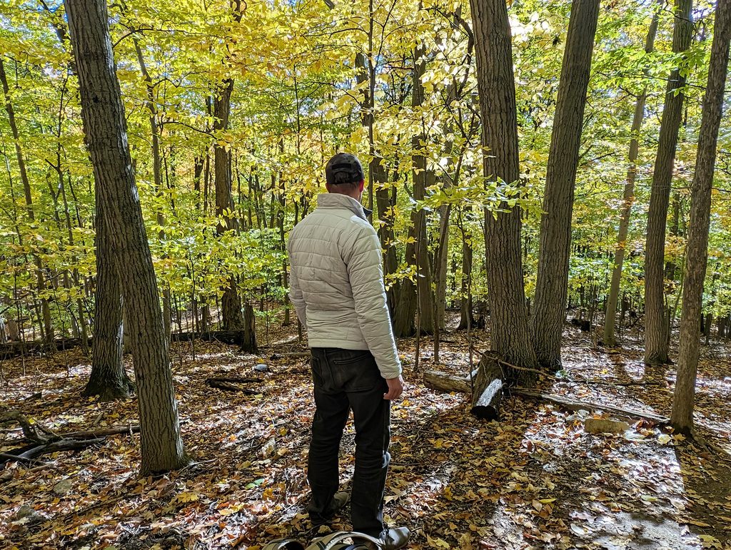 Helly Hansen Men's LIFALOFT™ Insulator Jacket Review: Three Weeks In, One Layering Lesson 5 Man in a beige jacket and black pants standing in a forest with autumn leaves and tall trees.
