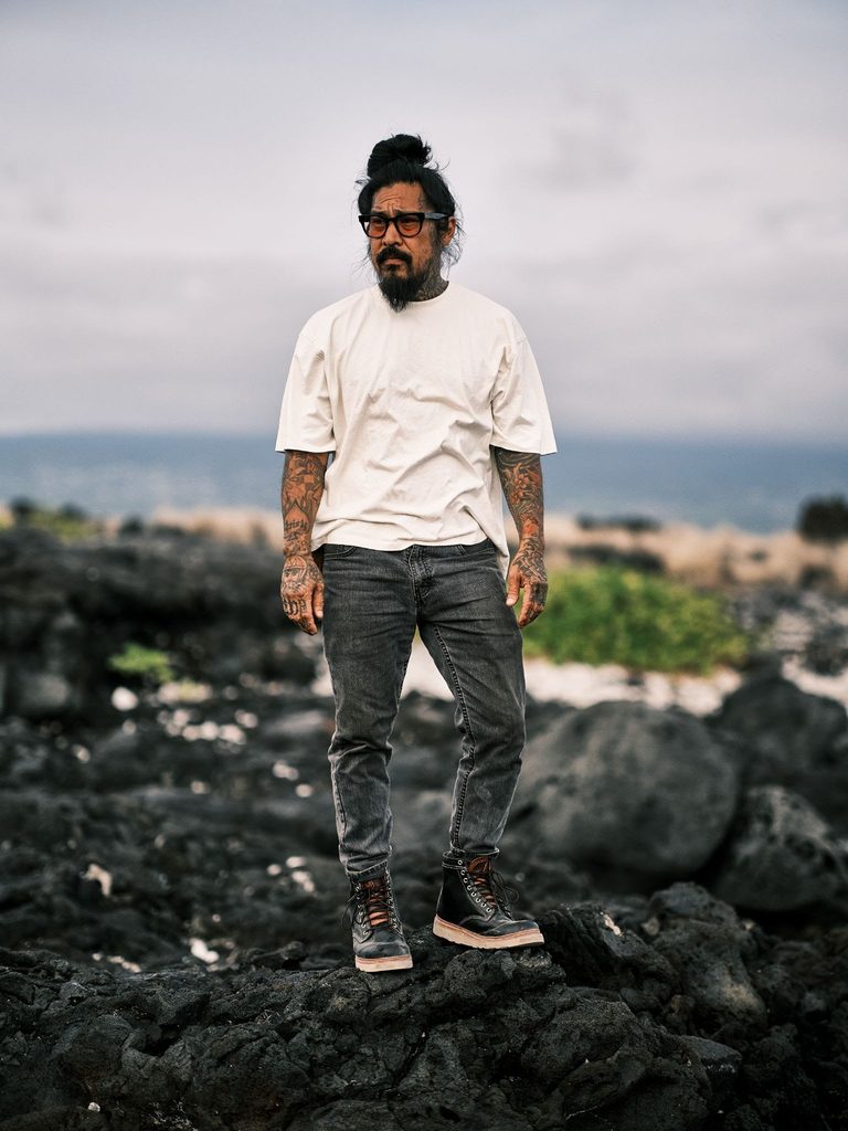 OluKai x Neil Kamimura Limited Edition 'Pāhi' Boots: Price, Release Date, and the Bladesmith Behind the Drop 2 Man with tattoos and glasses standing on volcanic rocks near the coast, wearing a white t-shirt and dark jeans.