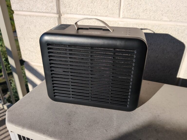 Black PuroAir 100i HEPA smart air purifier with handle, placed on a concrete surface outdoors, casting a shadow on the wall behind.