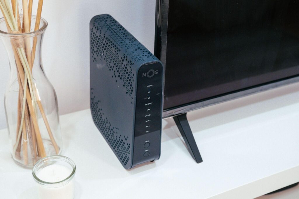 Modern WiFi router with LED indicators on a white desk next to a TV and a glass jar with wooden sticks.