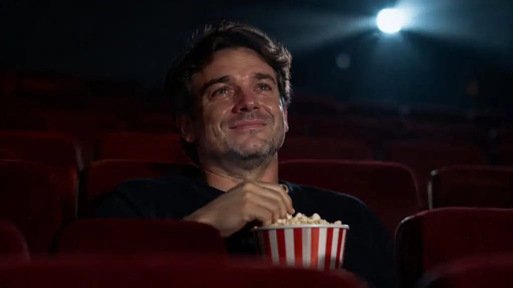 A man watching a movie in a dark cinema theater, holding a bucket of popcorn, with a joyful expression on his face.